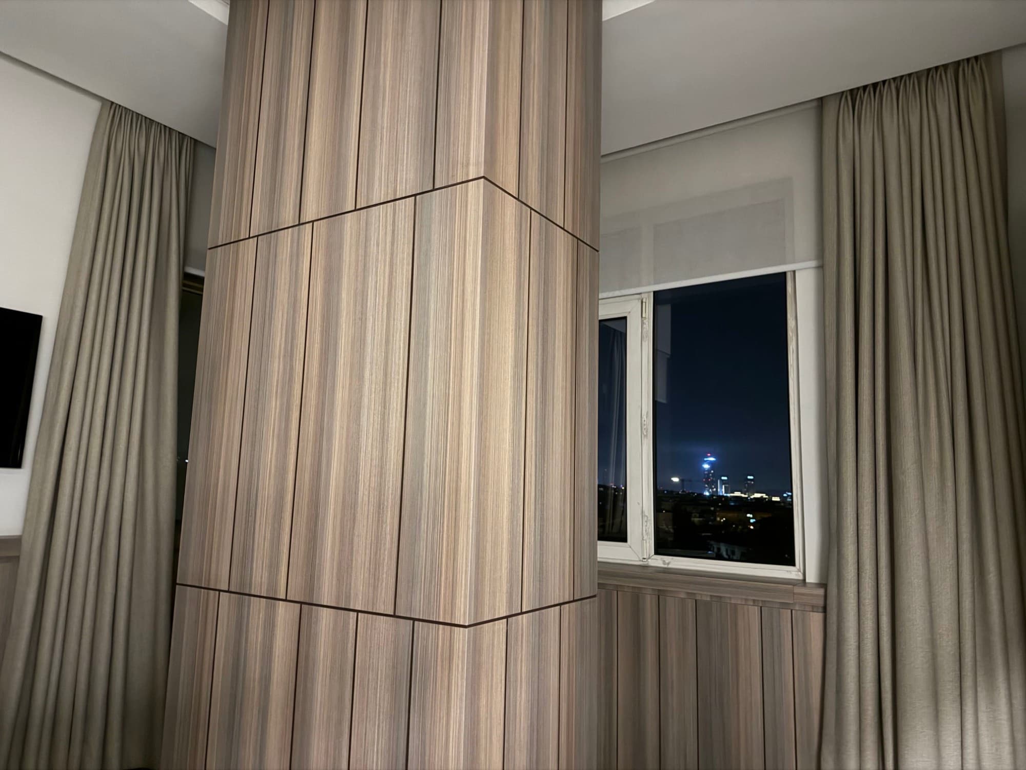 A wood-paneled room with floor-length curtains and a window looking out on the city at night.
