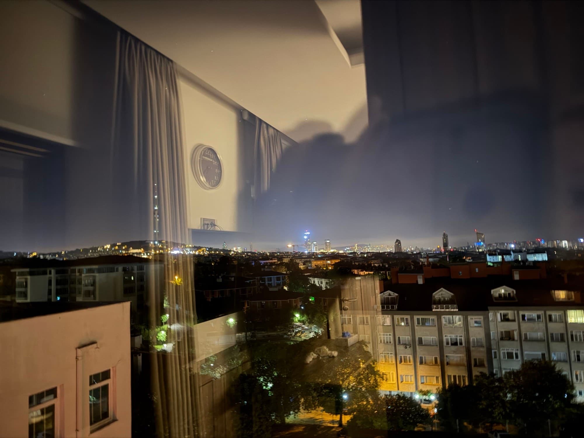 The Istanbul skyline at night, seen through a window with the room reflected in the glass.