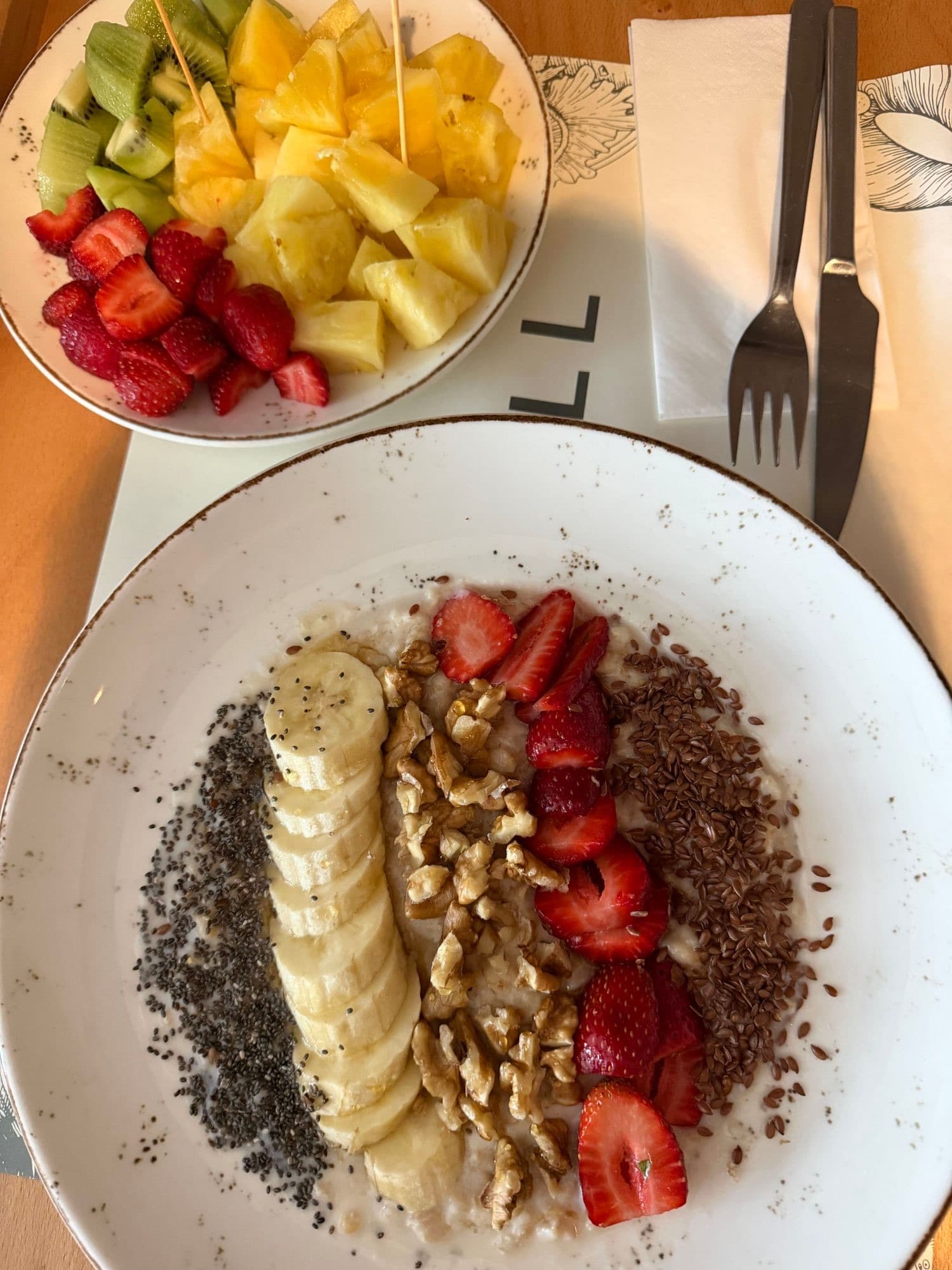Oatmeal topped with banana, walnuts, strawberries, chia, and flax seeds, with a bowl of fresh fruit.